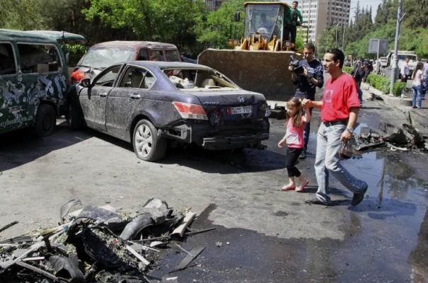 En man leder sin dotter förbi vraket av ett fordon efter en explosion i Mazzehdistriktet i Syriens huvudstad Damaskus den 29 april 2013, vilket tros ha varit riktat mot premiärministerns konvoj, enligt rapporter från Syriens statliga television.  (Foto: Louai Beshara/ AFP)
