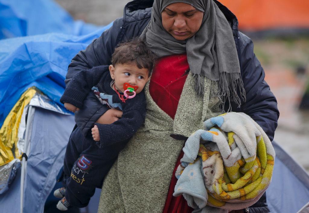 En kvinna bär en babys och en filt i det leriga tältlägret utanför Idomeni, där det fallit mycket regn. (Foto: Vadim Ghirda/AP/TT)