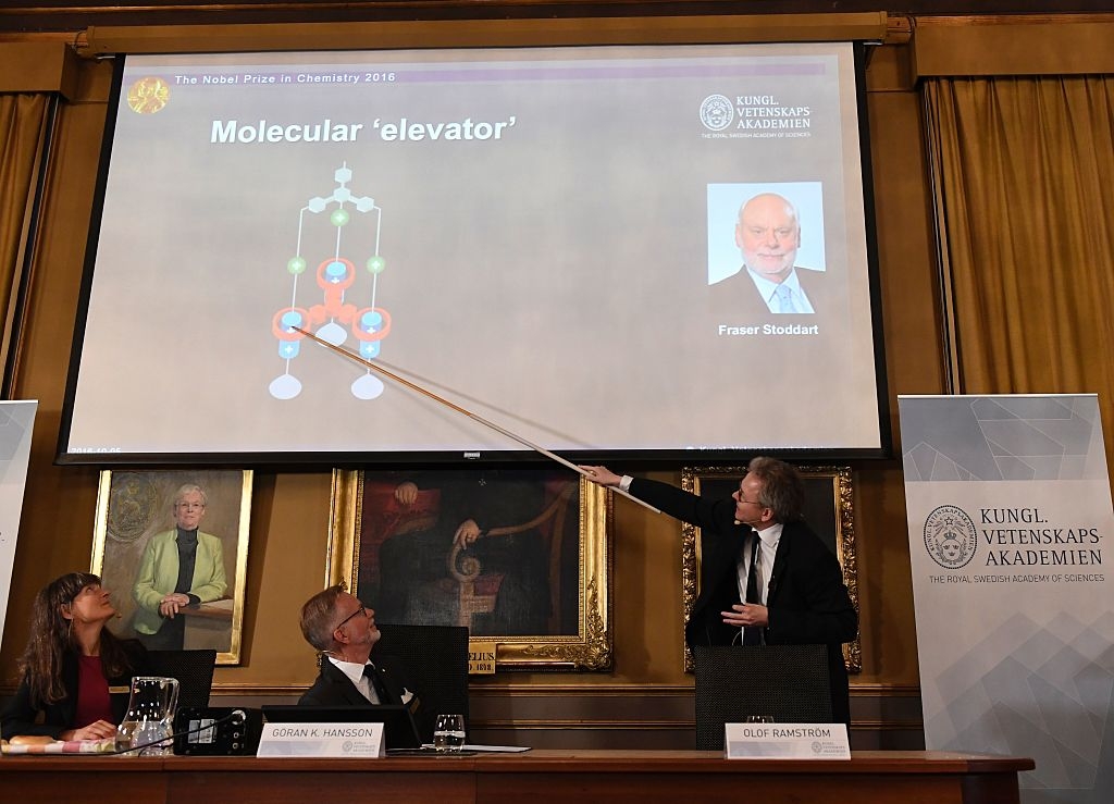 Professor Olof Ramström talar om den molekylära "hissen". Lyssnar gör ständige sekreteraren Göran K Hansson och professor  Sara Snogerup Linse, vid presskonferensen. På skärmen en av Nobelpristagarna: Fraser Stoddart. (Foto: Jonathan Nackstrand /AFP/Getty Images)