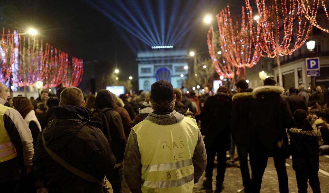 Demonstranter från Gula västarna tittar på ljusspelet över Triumfbågen i centrala Paris på nyårsafton. Foto: Kamil Zihnioglu/AP/TT