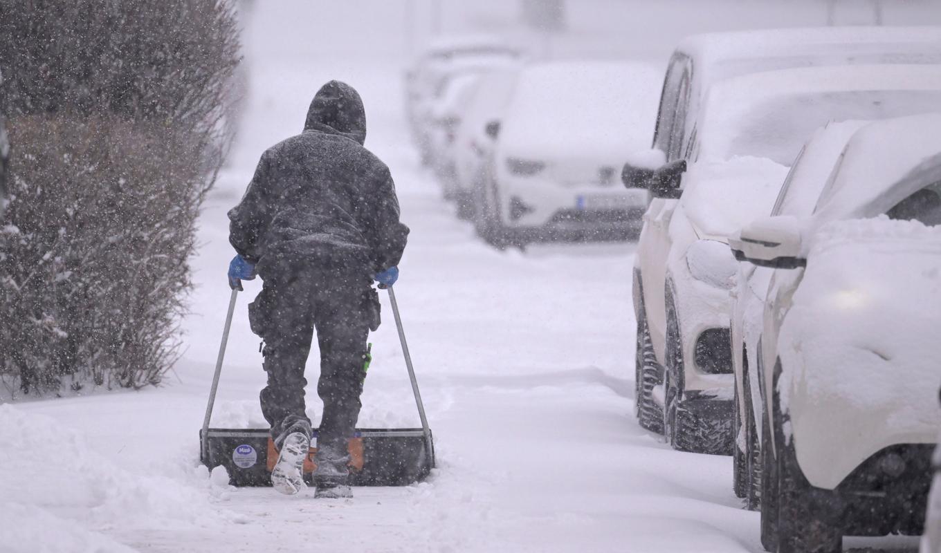 En man tar bort snö med en snöskovel på trottoaren mellan ett bostadshus och snötäckta parkerade bilar. Bild från november. Foto: Janerik Henriksson/TT