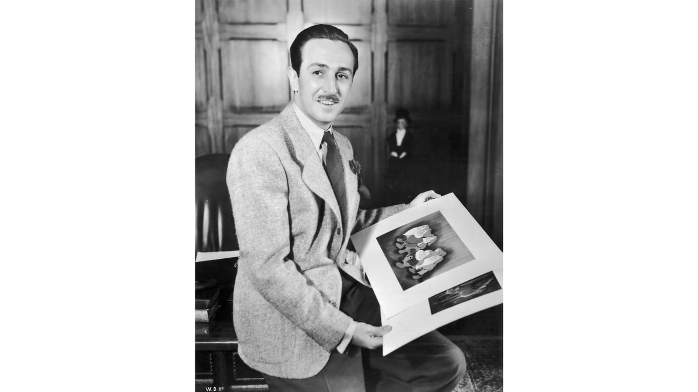 Cirka 1942. Walt Disney sitter på kanten på skrivbordet på sitt kontor och håller illustrationer från de animerade filmerna ”Snövit och de sju dvärgarna” och ”Bambi”. Den förstnämnda filmen gjorde att Walt kom på kant med sin fru. Foto: Hulton Archive/Getty Images