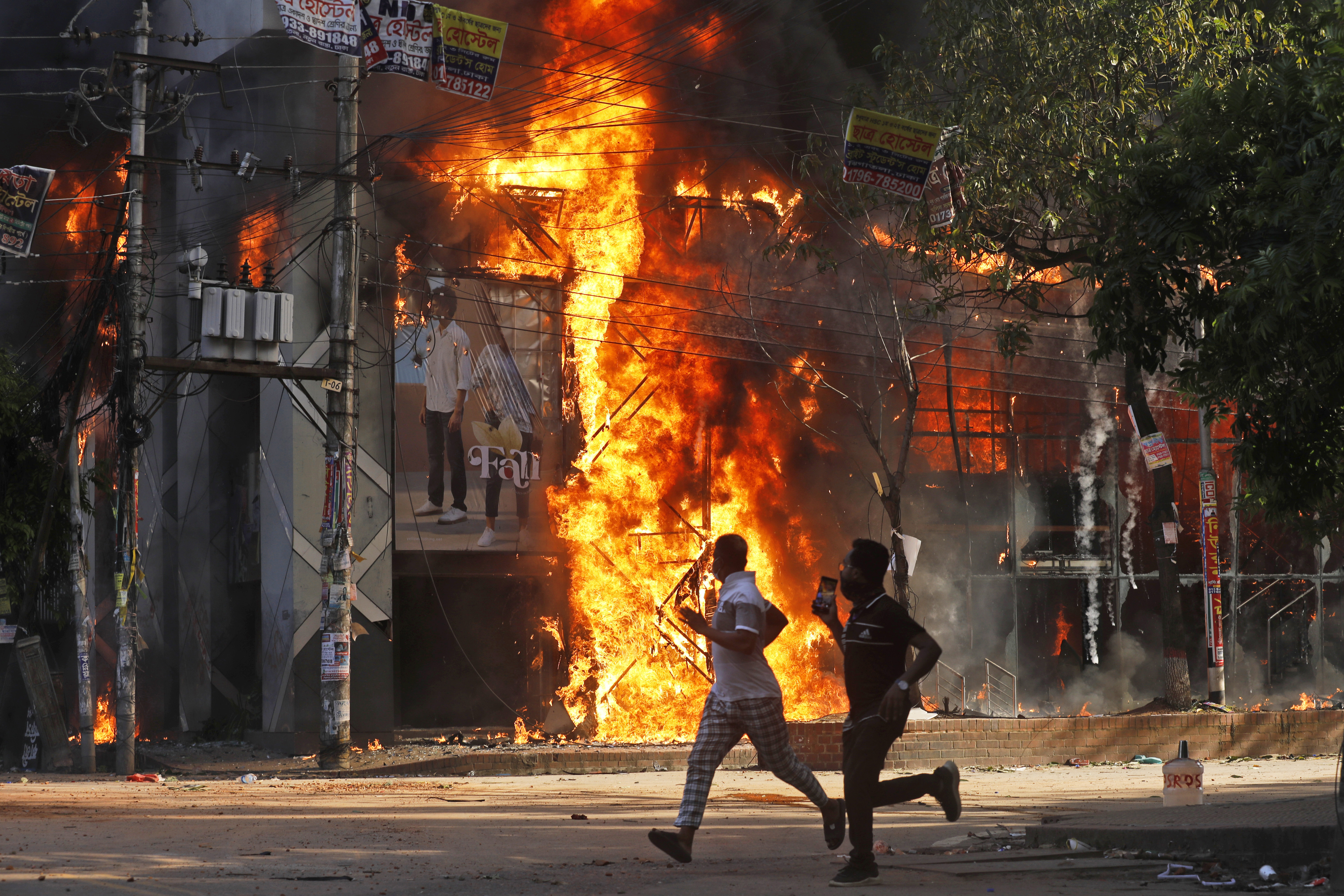 Två män springer under söndagen förbi ett köpcentrum som satts i brand under protesterna i Bangladesh. Foto: Rajib Dhar/AP/TT